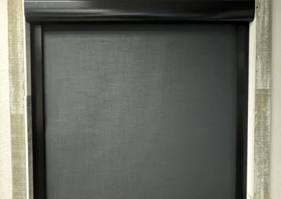 A black roller shade covers a window, framed by a rustic wooden border and a sturdy wooden sill.