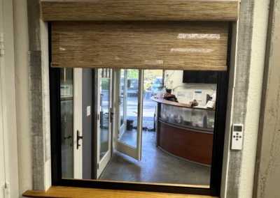 A modern window with a rolled bamboo shade, framed in wood and fabric, revealing a glimpse of an office interior.
