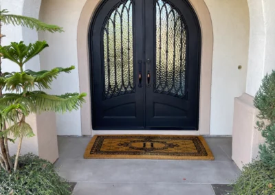Elegant black double doors with intricate glass designs, framed by greenery and a decorative welcome mat on a light-colored entryway.